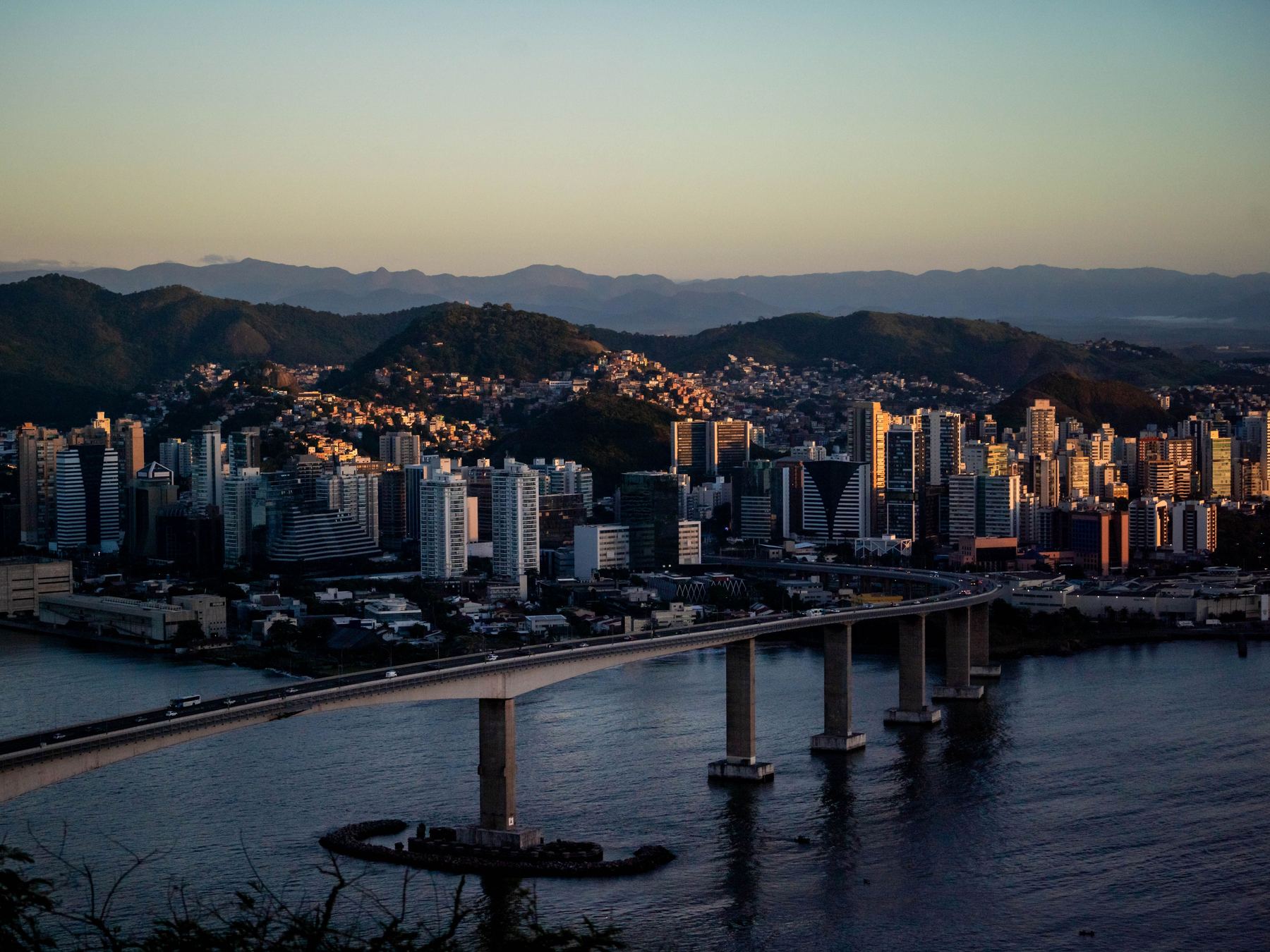 Terceira Ponte cruzando a baía no fim da tarde, vista de um morro de Vila Velha, com os prédios de Vitória iluminados pelo sol baixo e as montanhas no horizonte.