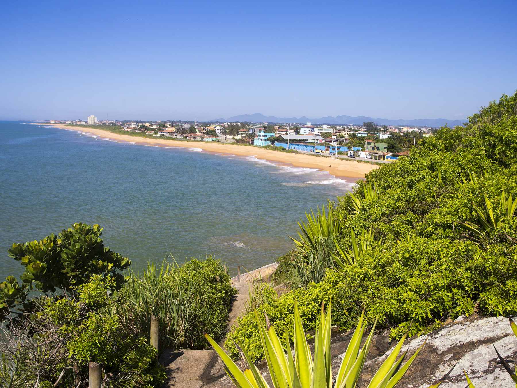 Praia da Barra do Jucu vista do alto do costão, com a faixa de areia curvando até as casas coloridas da vila e a vegetação de restinga em primeiro plano.