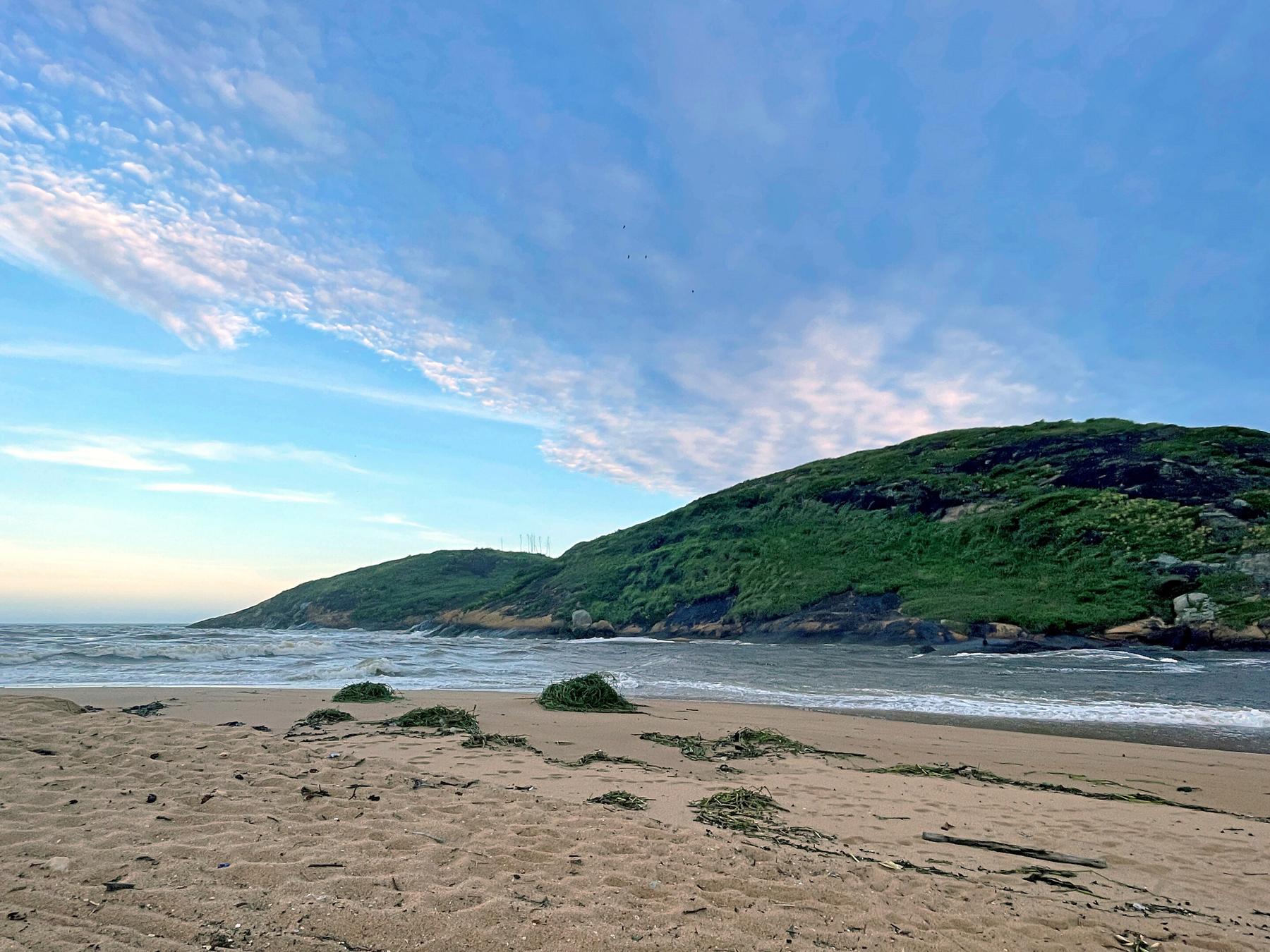 Foz do Rio Jucu encontrando o mar em manhã de céu claro, com a praia deserta em primeiro plano e o Morro da Concha coberto de vegetação verde à direita.