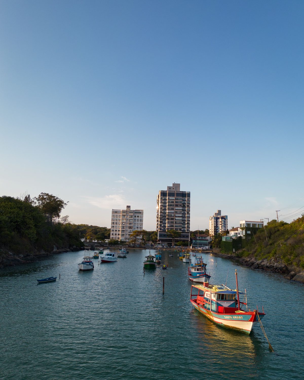 Barcos de pesca coloridos ancorados no canal da Praia da Costa ao fim da tarde, com prédios residenciais e vegetação nas duas margens.