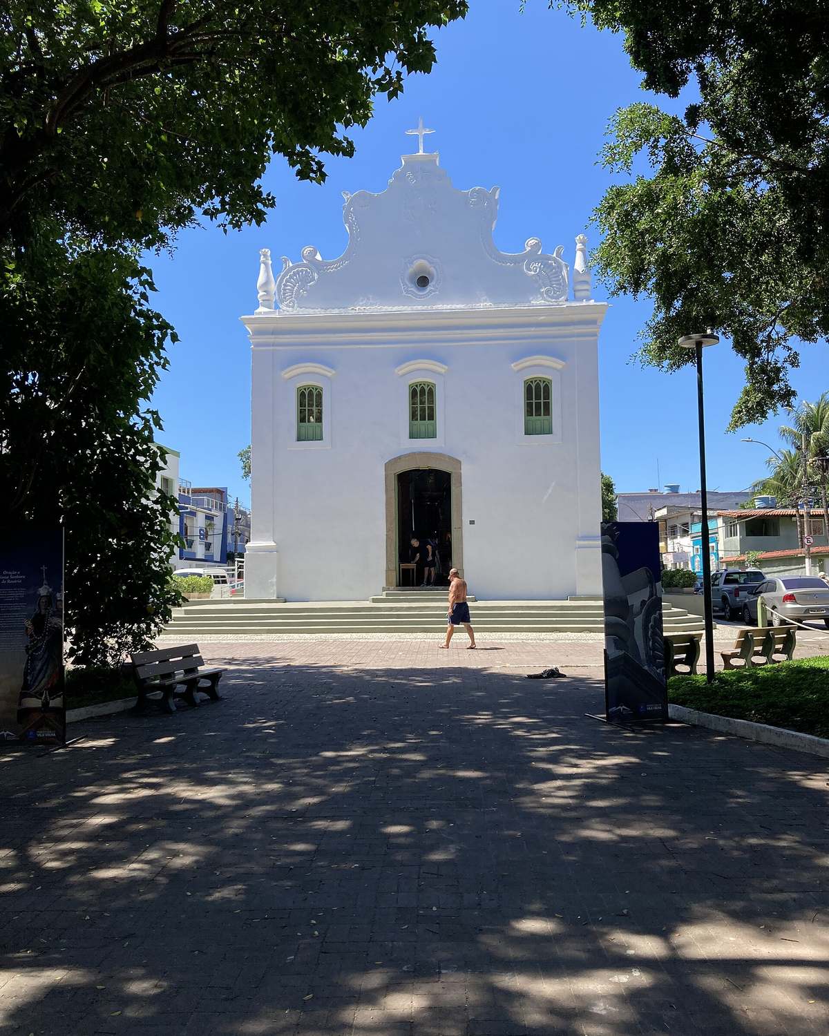 Fachada branca da Igreja de Nossa Senhora do Rosário, na Prainha, vista do largo arborizado, com uma pessoa passando na frente da escadaria.