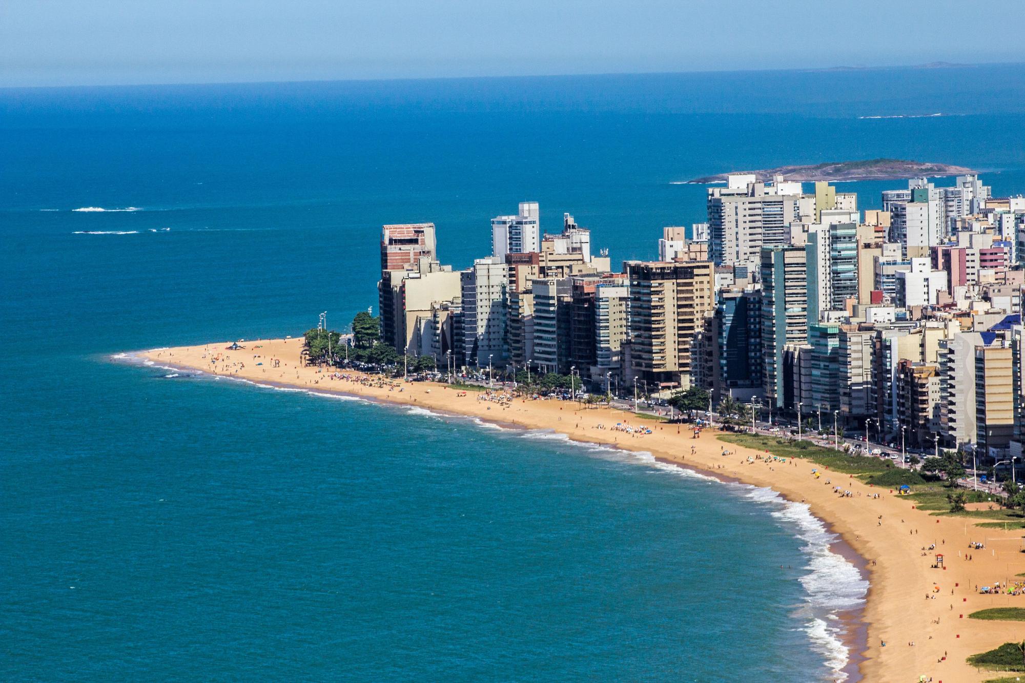Orla de Vila Velha vista do alto, com a faixa de areia curvando ao lado dos prédios altos da praia, banhistas espalhados pela areia e o mar azul em primeiro plano.