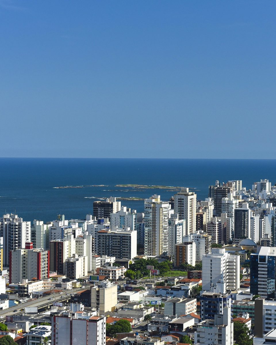 Vista aérea da orla verticalizada de Vila Velha, com dezenas de prédios altos colados uns nos outros, o mar azul e as pedras da arrebentação ao fundo.