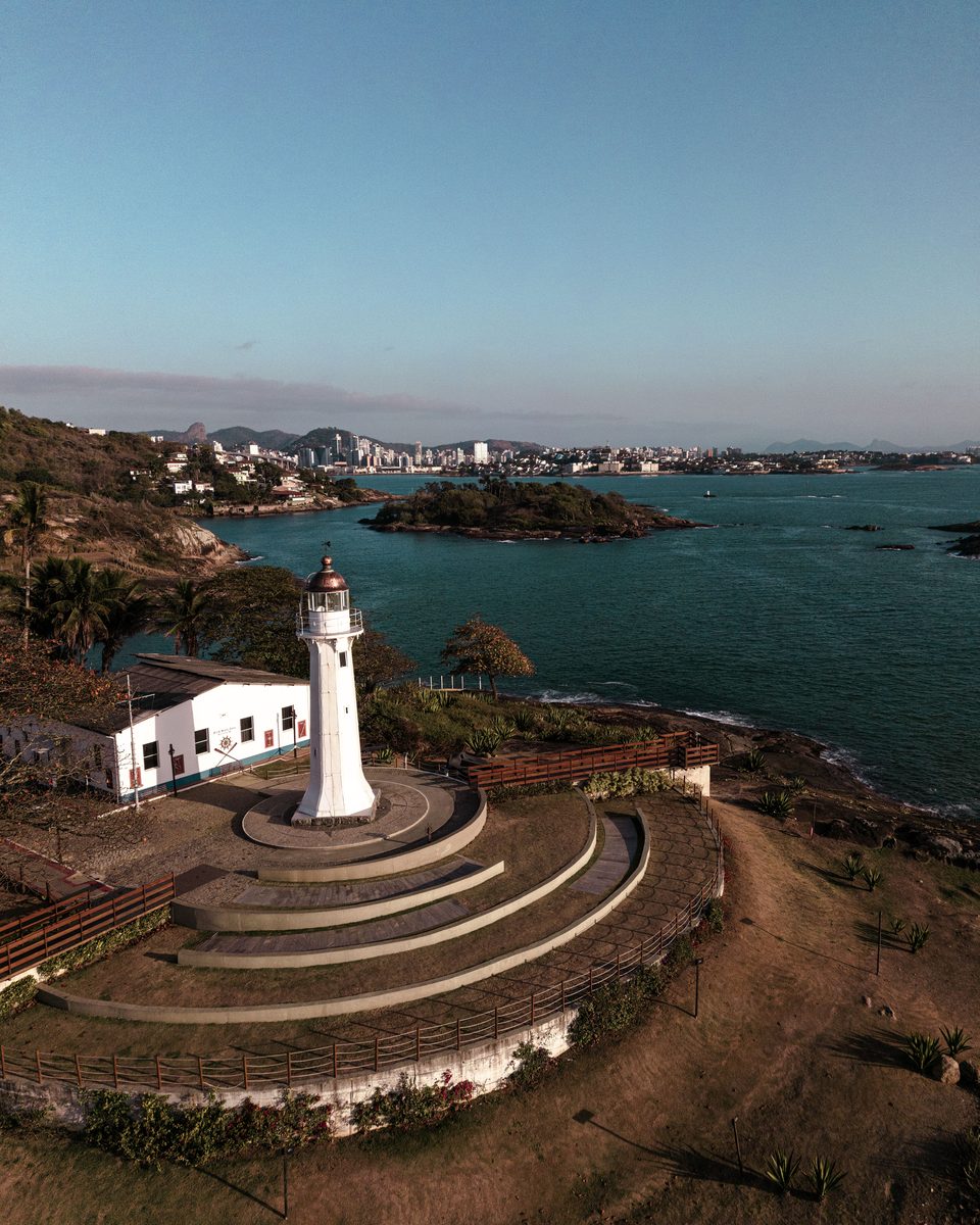 Farol branco de Santa Luzia sobre a ponta de pedra ao lado do mar, com a casa do faroleiro, o mirante em degraus e a cidade ao fundo do outro lado da água.