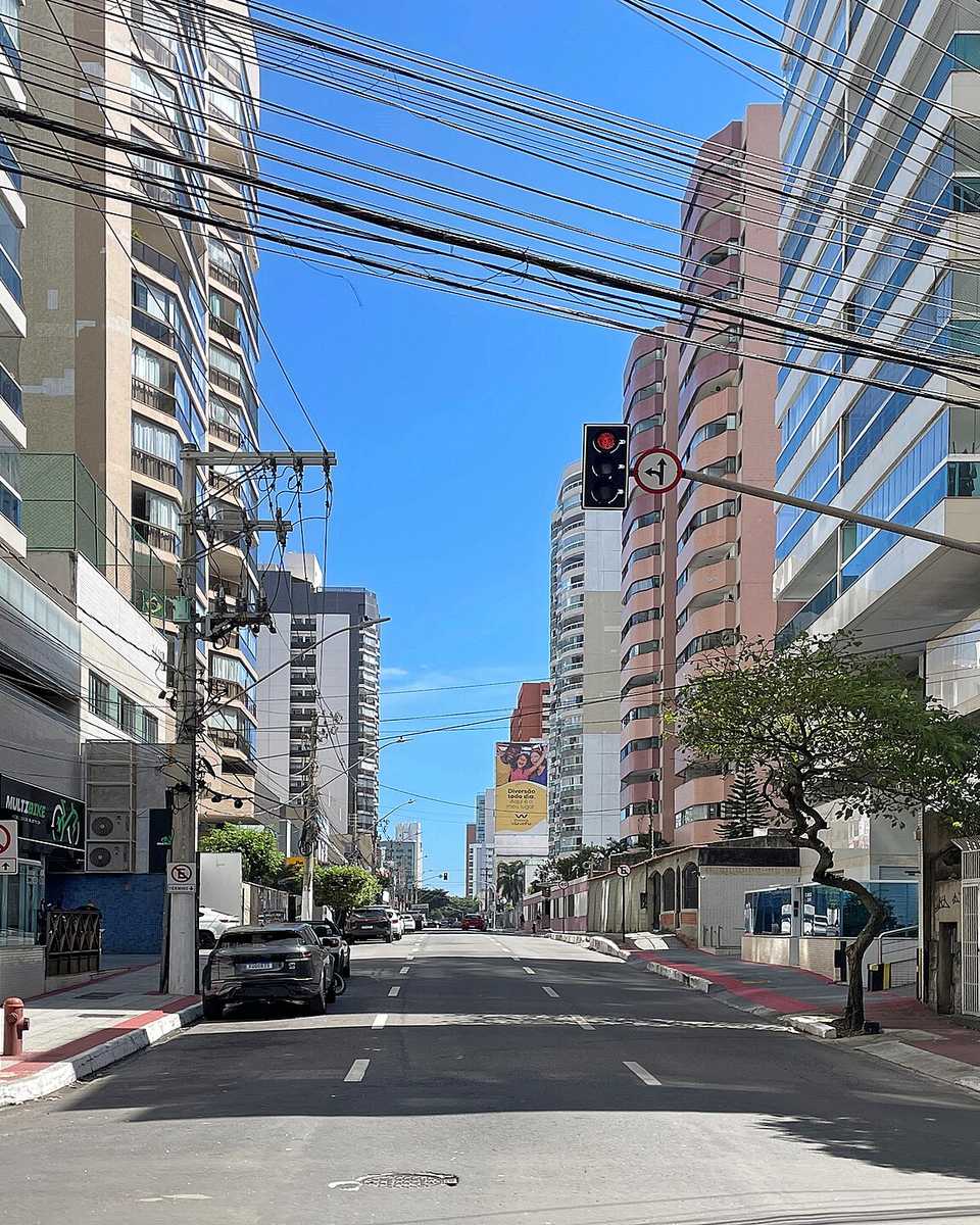 Rua do bairro de Itapoã com prédios residenciais dos dois lados, lojas no térreo, semáforo, bicicletas de aluguel na calçada e o céu azul ao fundo.
