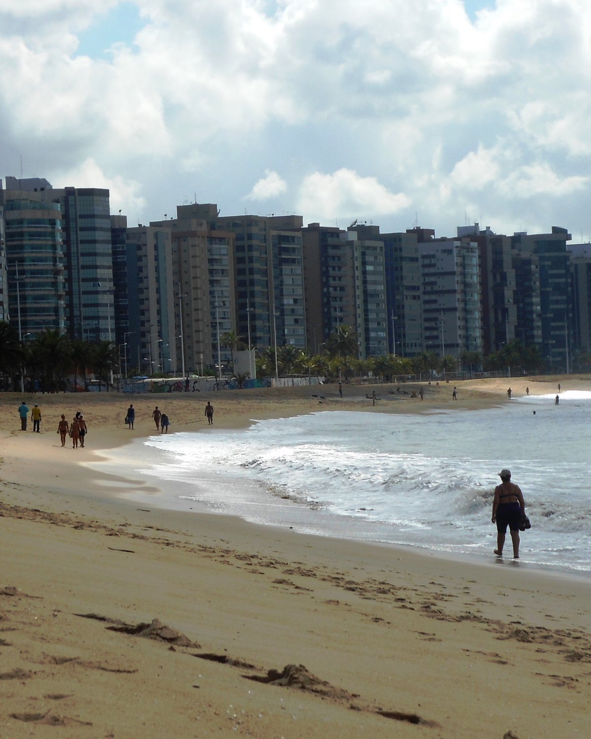 Pessoas caminhando na areia da Praia de Camburi em fim de tarde, com a fileira de prédios da orla e o calçadão ao fundo.
