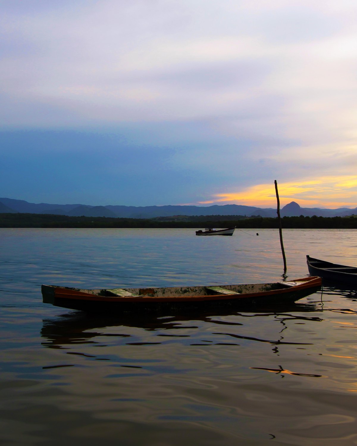 Canoas de madeira paradas na água calma da baía no fim da tarde, na Ilha das Caieiras, com o morro recortado no horizonte e o céu dourado.