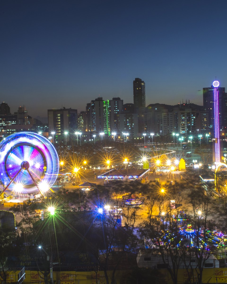 Parque de diversões iluminado à noite na Enseada do Suá, com roda-gigante colorida e brinquedos acesos, e os prédios de Vitória ao fundo.