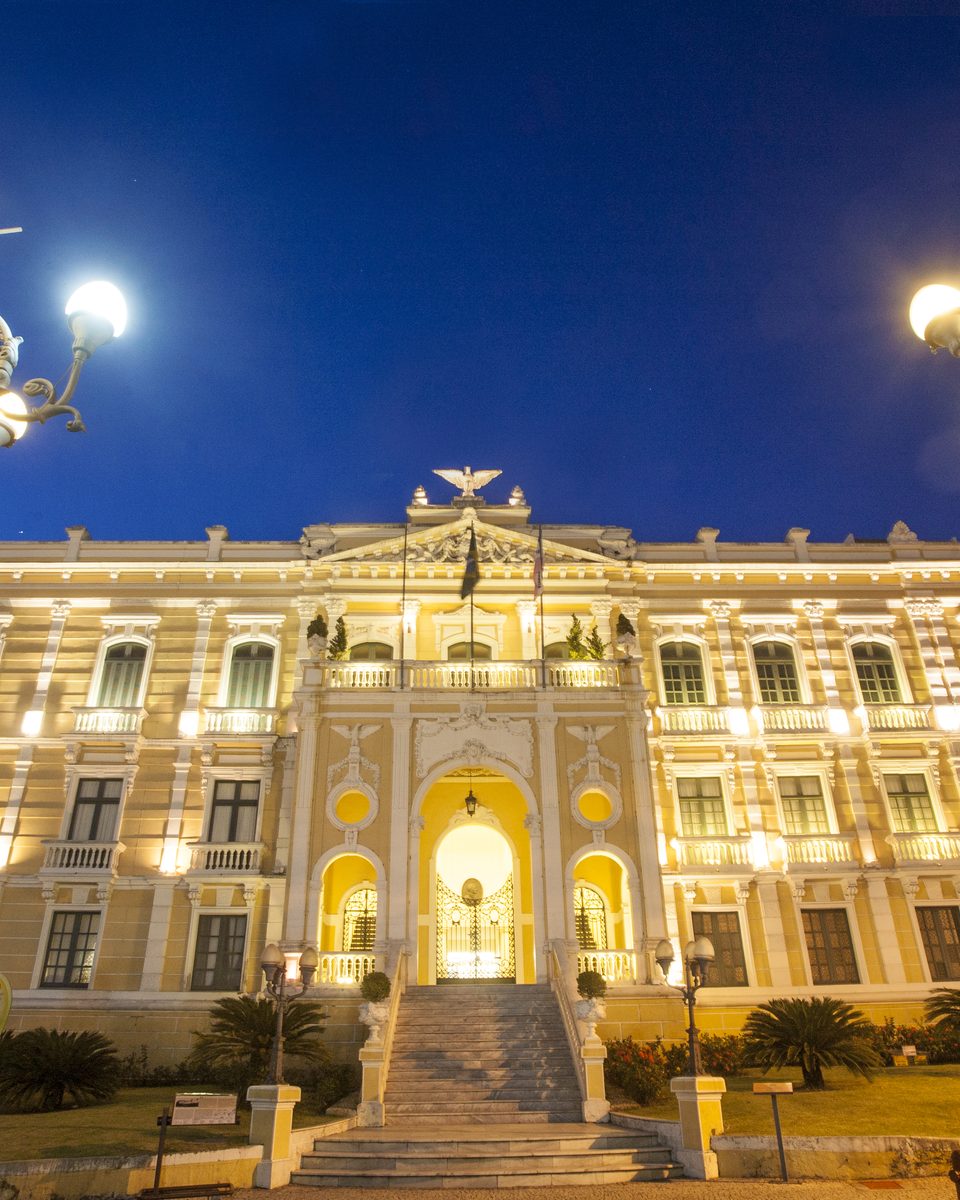 Palácio Anchieta iluminado ao anoitecer, com a fachada amarela e branca, a escadaria de acesso e dois lampiões antigos de ferro em primeiro plano.