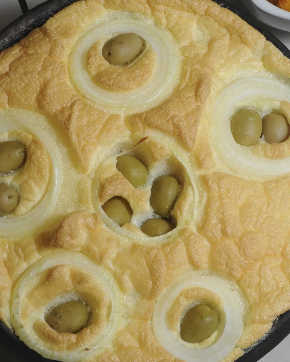 Torta capixaba assada na panela de barro preta, com rodelas de cebola e azeitonas por cima, servida sobre a mesa com arroz e farofa ao lado.