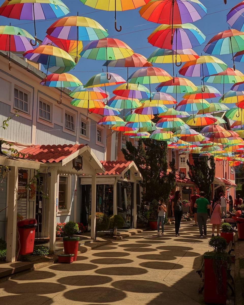 Rua de lojas em Penedo, distrito de Itatiaia, com casas de madeira coloridas, guarda-chuvas coloridos pendurados sobre a passagem e pessoas passeando num dia de sol.